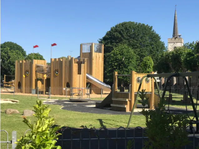 Completed timber playground at Newmarket Memorial Gardens with slides, swings, mature trees and the church spire beyond