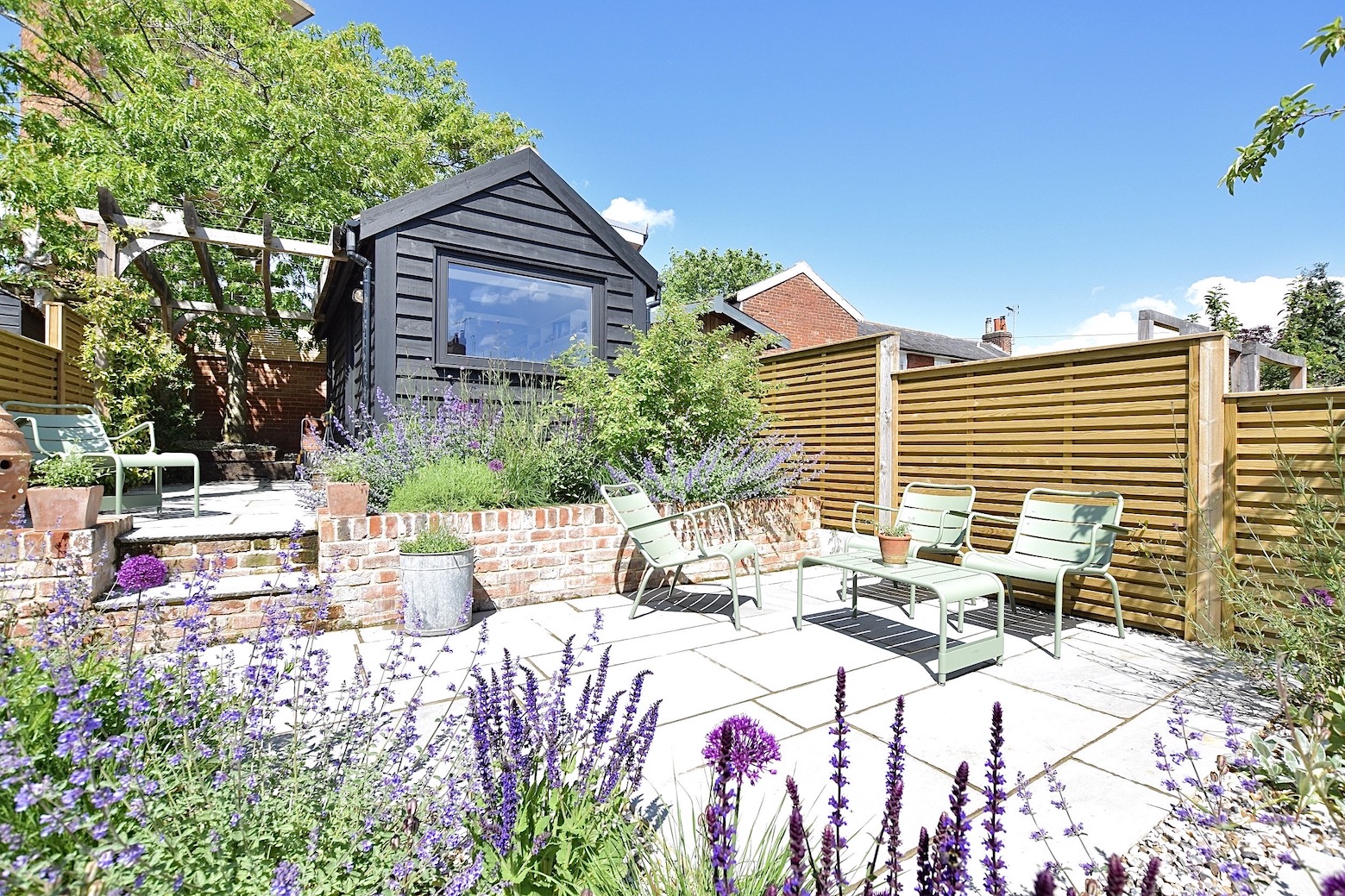 Upper seating terrace with pale paving, brick retaining walls, planting and dark garden office