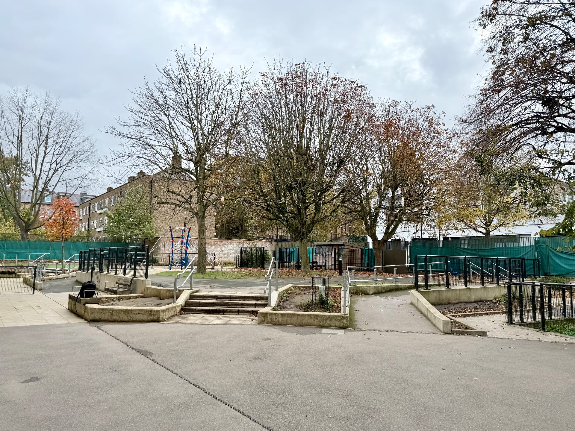 The existing school grounds in autumn, with foreground paving, raised beds, steps, mature trees, the school building and perimeter fencing