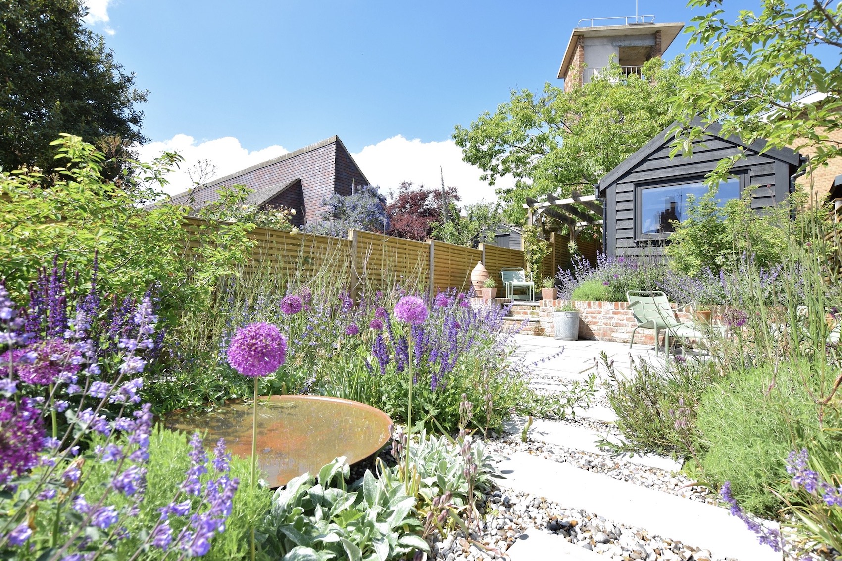 Richly planted Woodbridge garden with a pale path leading toward a dark garden office